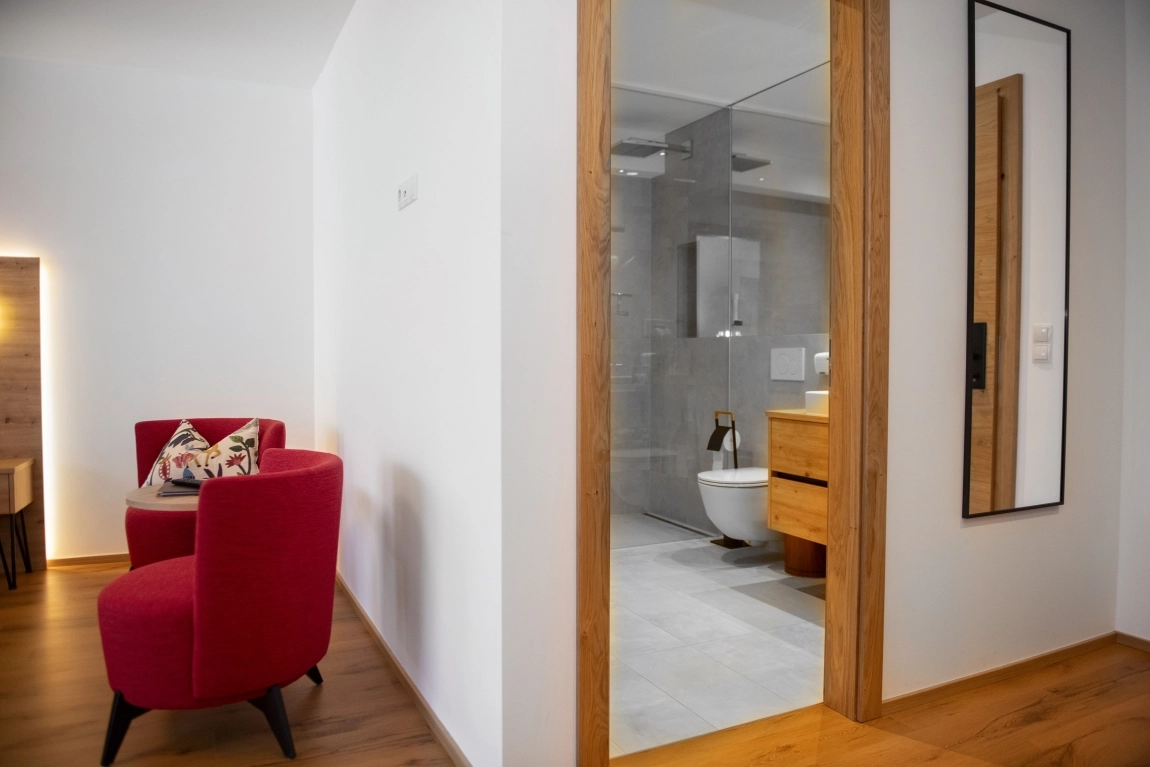 Blick vom Zimmer ins moderne Bad mit Glasdusche, WC und Waschtisch, daneben rote Sessel und weißer Wandspiegel.