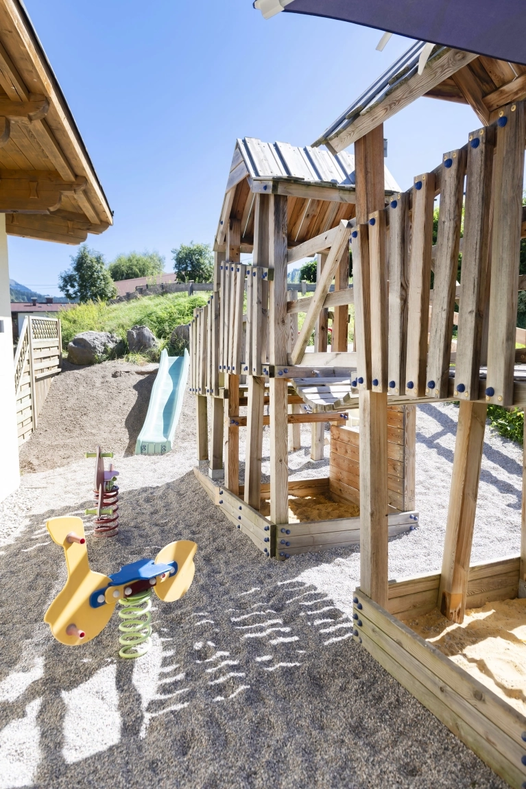 Spielplatz mit Holzkletterturm, Rutsche, Federwippe und Kiesboden im sonnigen Außenbereich des Hotels.