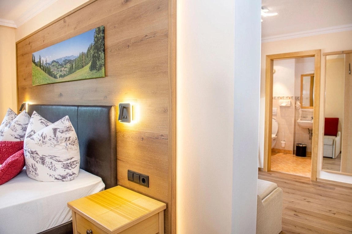 Linke Betthälfte mit Naturmotiv-Kissen, daneben Nachttisch; rechts Tür zum hell gefliesten Badezimmer.