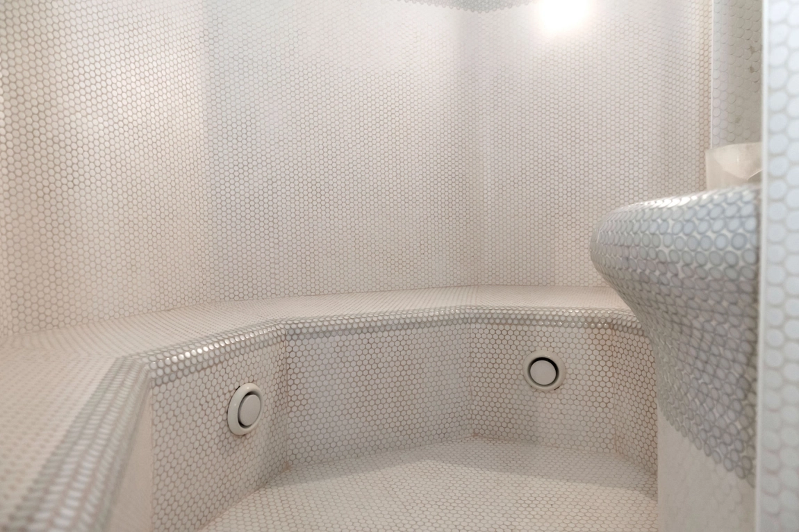 Heller Dampfbadraum mit runder Sitzbank, kleinteiligen Mosaikfliesen, Dampföffnung und weicher Lichtquelle zur Entspannung.