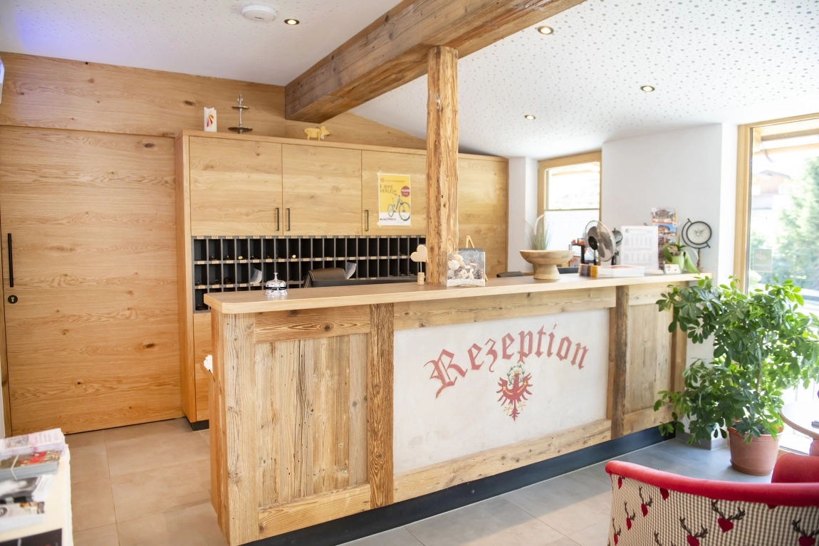 Helle Hotelrezeption mit Holztheke, Schriftzug ‚Rezeption‘, Schlüsselregal im Hintergrund und Pflanzen im Raum.