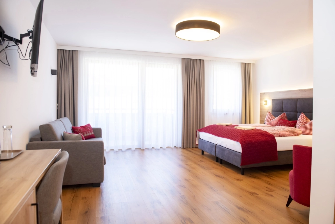 Großes, helles Zimmer mit Doppelbett, Sofa, Schreibtisch und TV. Holzfußboden, Vorhänge und viel Tageslicht.