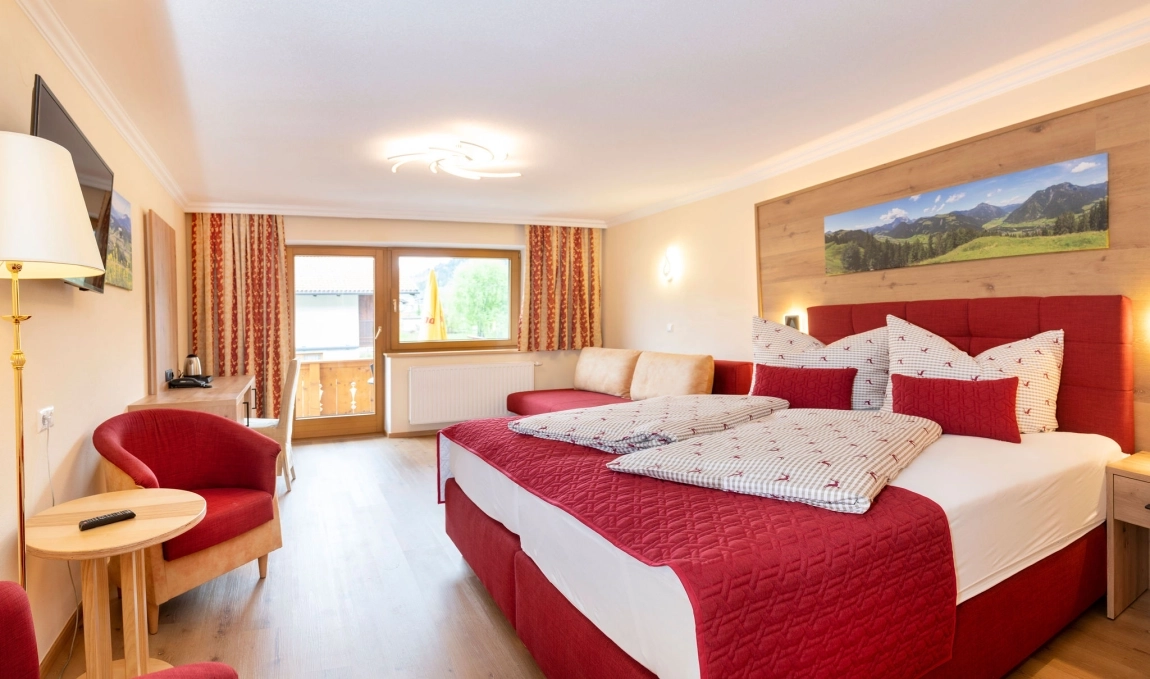 ChatGPT said: Helles Zimmer mit rotem Doppelbett, Sofa, Schreibtisch, zwei Sesseln und Balkon mit Blick ins Grüne.
