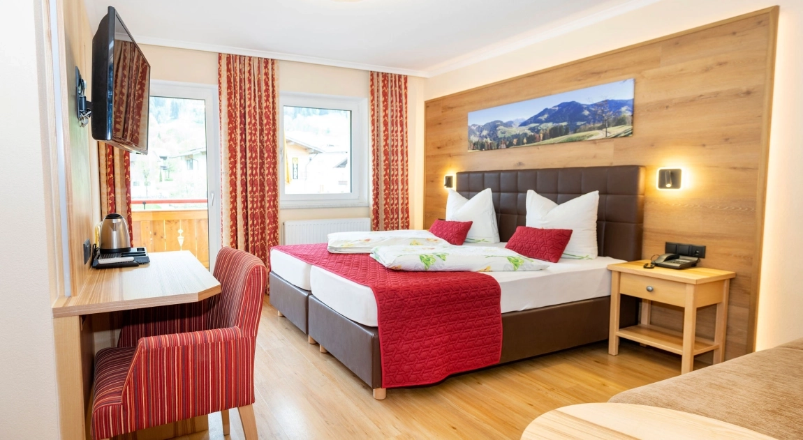Helles Hotelzimmer mit Doppelbett, Schreibtisch, TV, roten Vorhängen und großem Fenster mit Blick nach draußen.