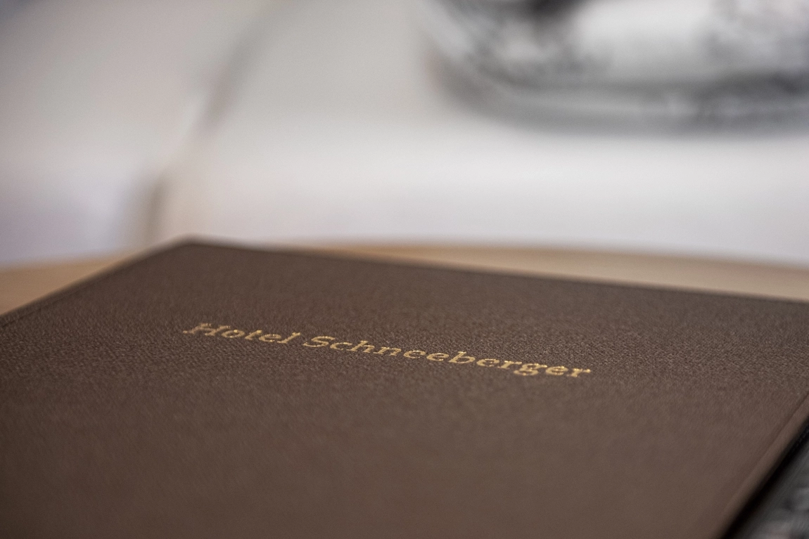 Nahaufnahme einer braunen Gästemappe mit goldener Schrift „Hotel Schneeberger“ auf einem Tisch im Zimmer.