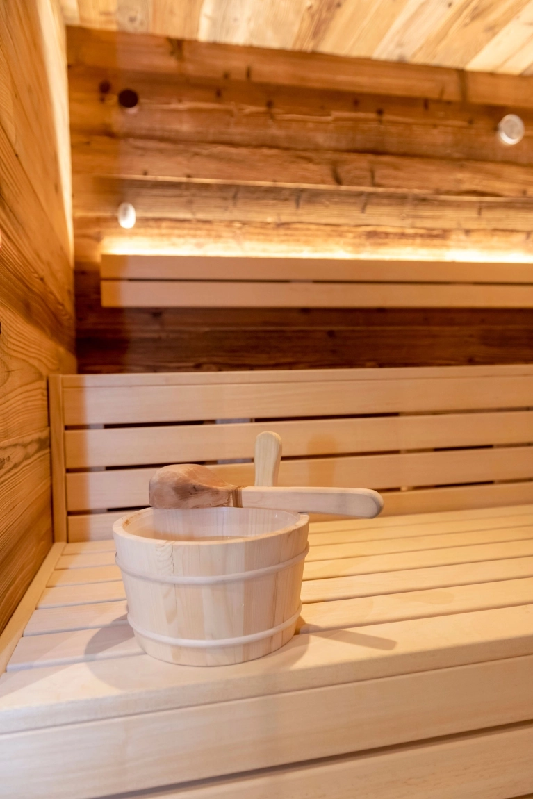 Ein Holz-Eimer mit einem Holzlöffel steht auf einer hölzernen Saunabank, umgeben von Holzwänden und sanfter Beleuchtung.