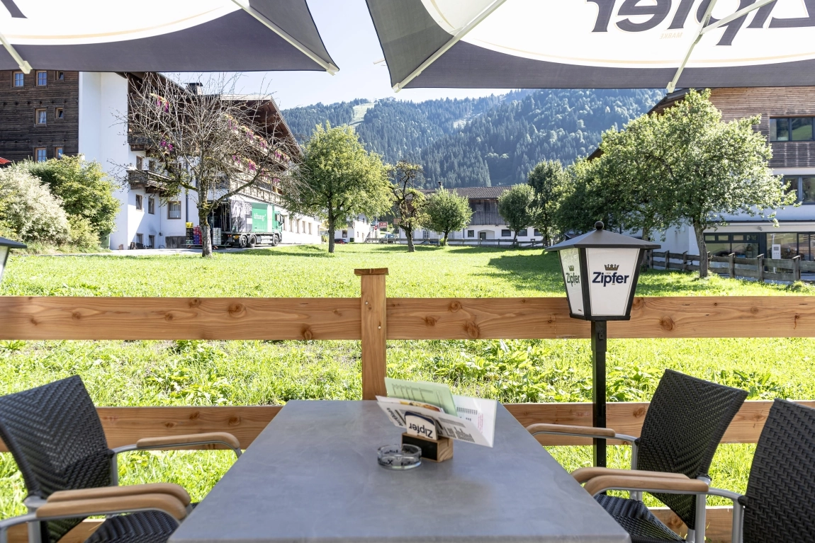Sitzplatz auf Hotelterrasse mit Blick auf Wiese, Holzzaun, umliegende Gebäude und bewaldete Hügel.