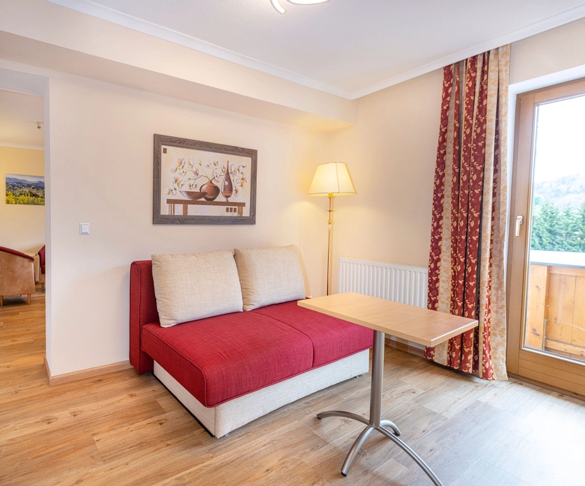 Rotes Sofa mit beigen Kissen, davor ein Holztisch, daneben Stehlampe und Tür zum Balkon mit Vorhängen.