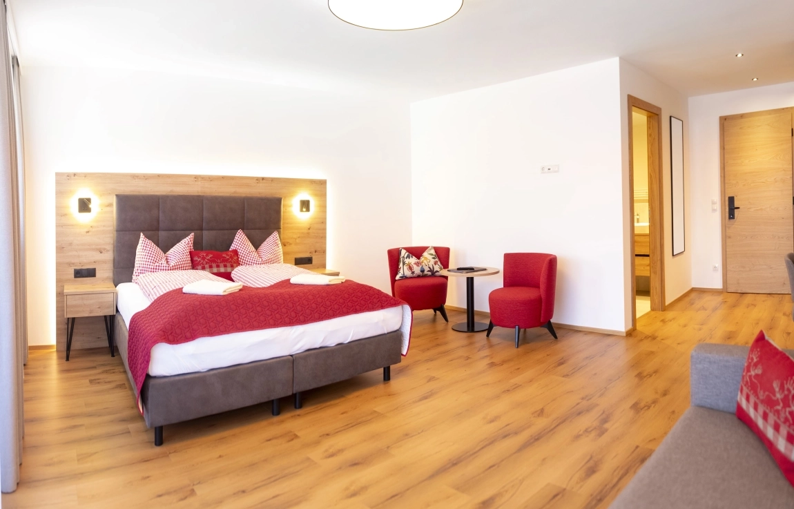 Helles Hotelzimmer mit Doppelbett, roten Akzenten, Sitzgruppe und Holzboden, rechts ein Badzugang.