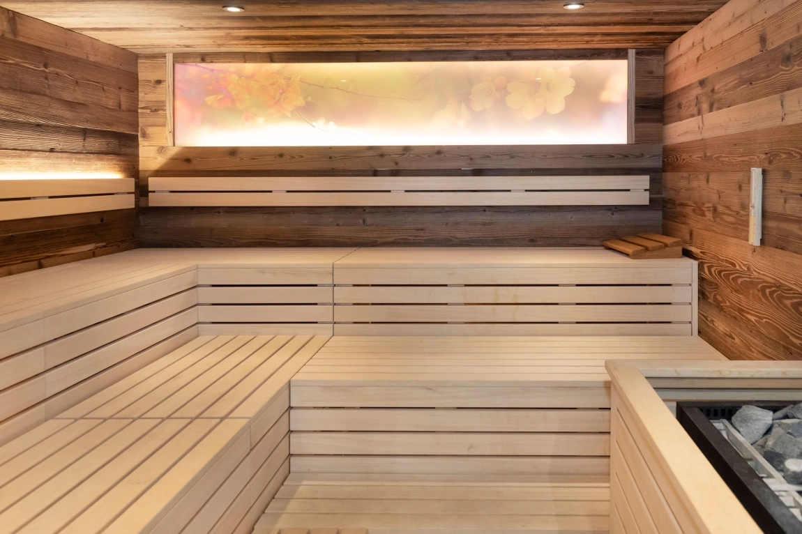 Innenaufnahme einer Sauna mit hellen Holzflächen, Bänken und einem beleuchteten Fenster, das sanftes, diffuses Licht abgibt.