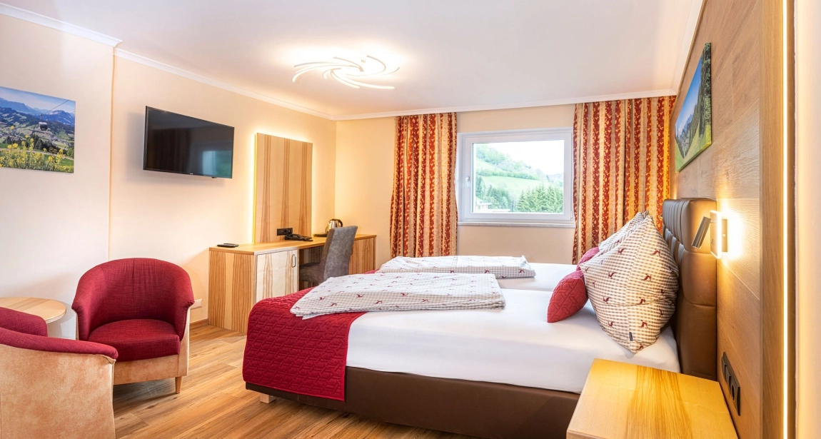 Hotelzimmer mit Doppelbett, Schreibtisch, TV, Sesseln und Fenster mit Blick ins Grüne, warm beleuchtet.
