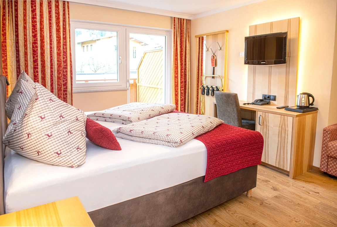 Helles Zimmer mit Doppelbett, rotem Dekor, Schreibtisch, TV an der Wand und Blick durch Fenster auf Holzverkleidung draußen.