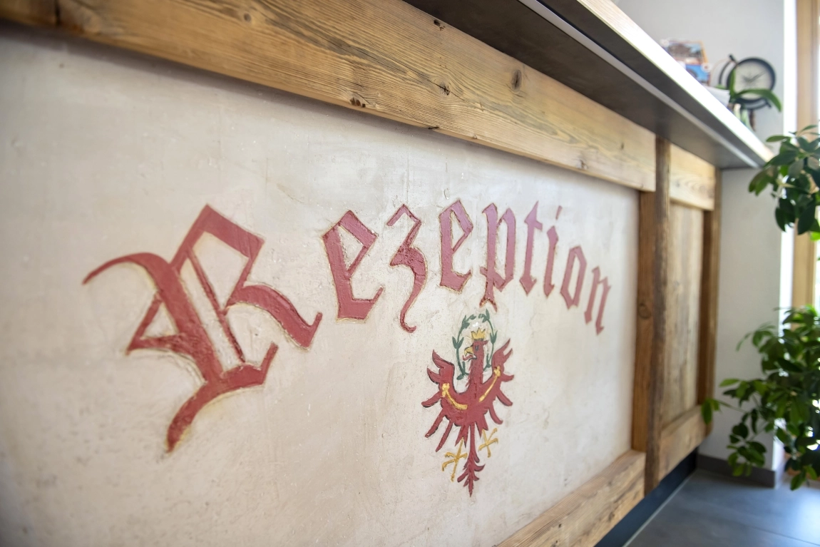 Holztheke mit dem Wort ‚Rezeption‘ in altdeutscher Schrift und Tiroler Adler-Emblem, Pflanze am rechten Bildrand.