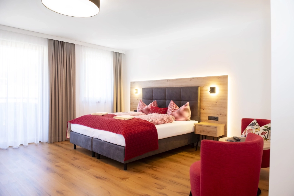 Modernes Zimmer mit Doppelbett, rotem Sessel, Nachttischen und viel Licht durch große Fenster mit Vorhängen.