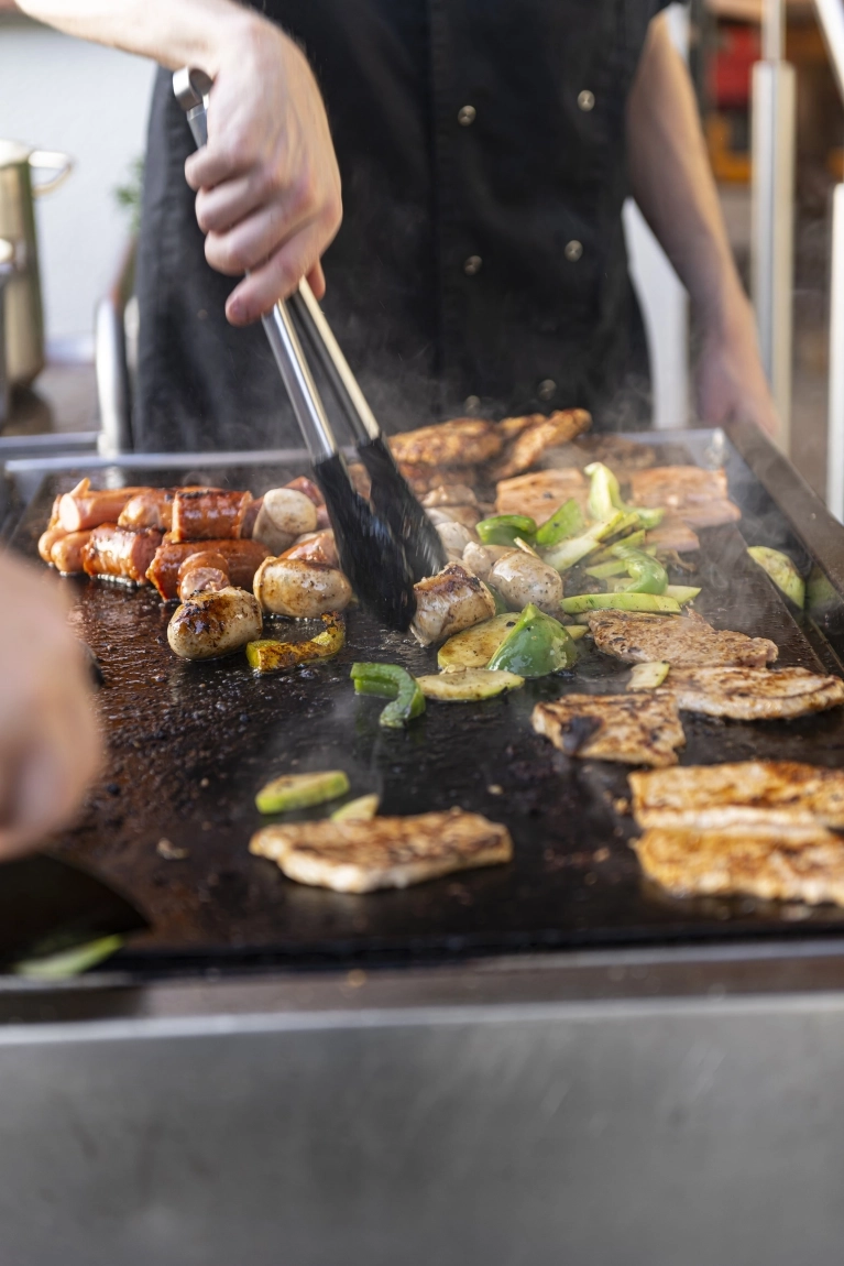 Person grillt Fleisch, Würstchen und Gemüse auf großer Grillplatte, dampfend und frisch zubereitet.