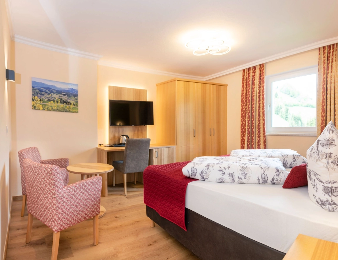 Helles Hotelzimmer mit Doppelbett, TV, Holzschrank, Fenster mit Ausblick und zwei Sesseln mit Tisch.