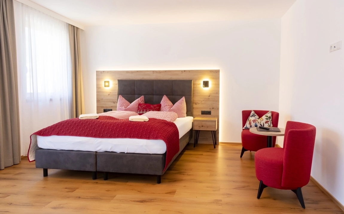 Doppelbett mit rot-weiß karierten Kissen, zwei roten Sesseln und viel Tageslicht durch ein Fenster links.