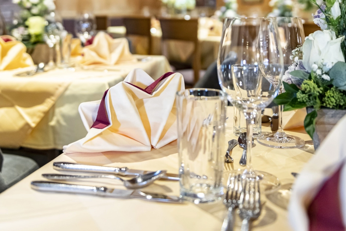 Nahaufnahme eines gedeckten Restauranttisches mit Besteck, Gläsern, gefalteter Serviette und Blumenschmuck.