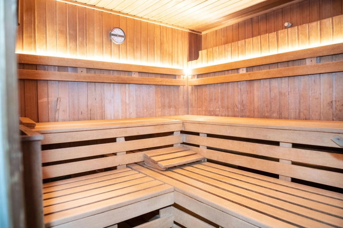 Helle Sauna aus Holz mit breiten Sitzflächen, Rückenlehnen, indirektem Licht und einem Thermometer an der Wand.