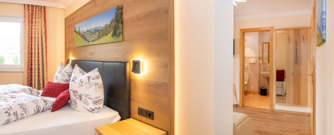 Helles Doppelzimmer mit Holzmöbeln, Bett mit Naturmotiv, rotem Kissen und Blick ins angrenzende Badezimmer.