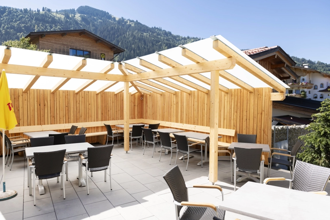 Sonnige Terrasse mit Überdachung, mehreren Sitzgruppen und Ausblick auf umliegende Hänge und Gebäude.