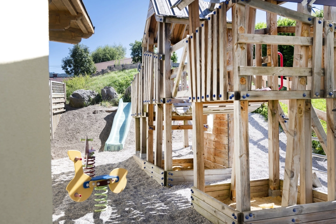 Kinderspielplatz mit Holzklettergerüst, Rutsche, Wippe und Kiesboden im Außenbereich nahe dem Hotel.