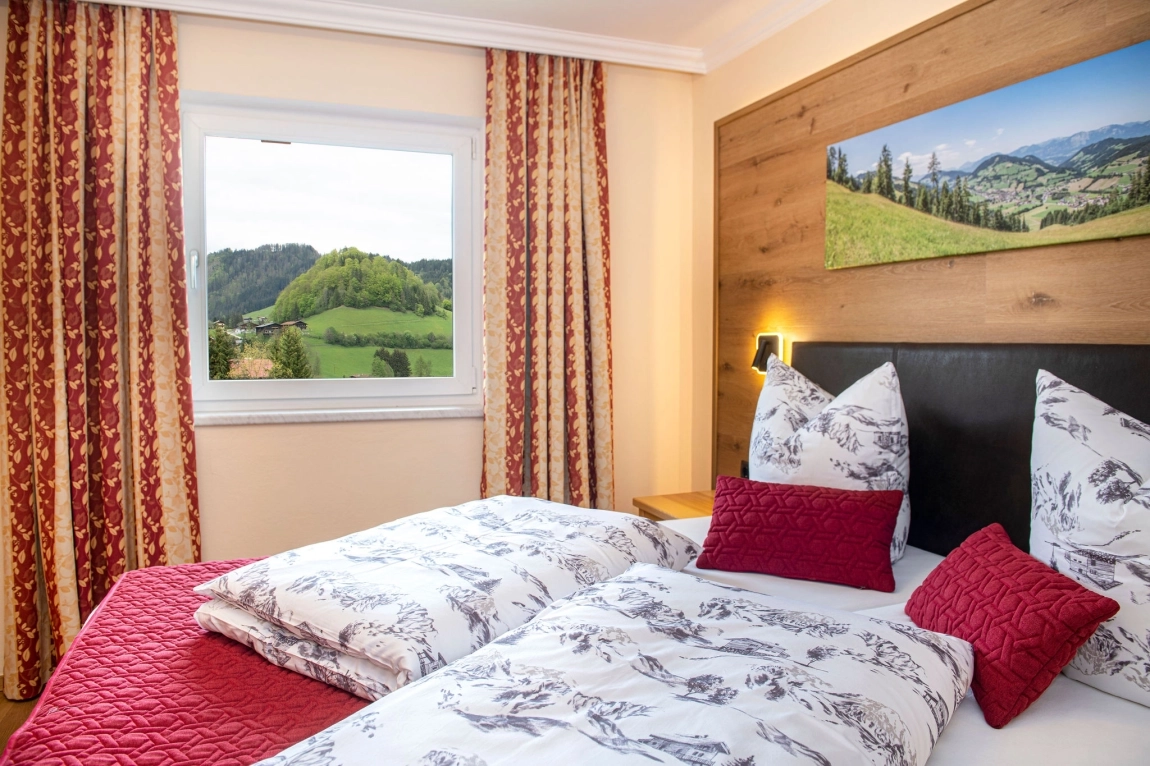 Doppelbett mit weiß-roter Bettwäsche vor Fenster mit Aussicht auf grüne Hügel und Waldlandschaft.