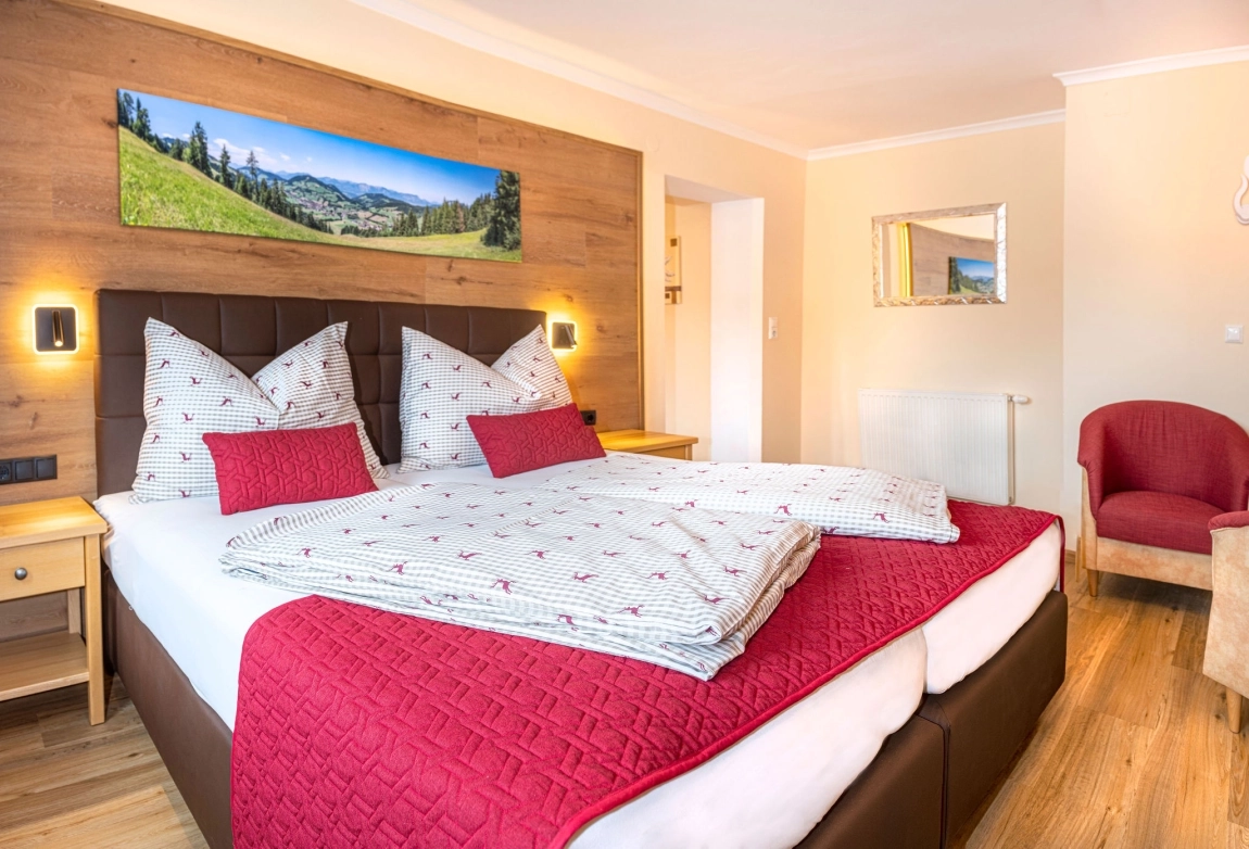 Doppelbett mit rot-weißer Bettwäsche in hellem Zimmer, darüber Panorama-Foto, Holzfußboden, gemütliche Atmosphäre.