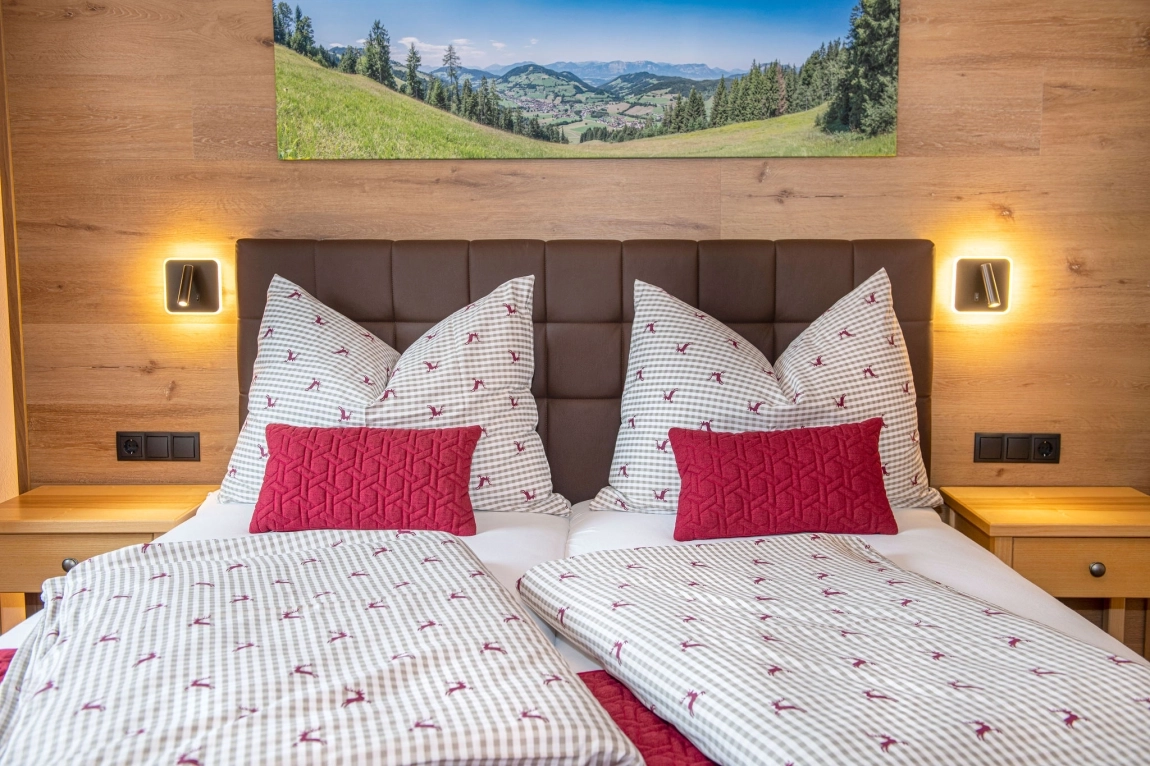 Doppelbett mit gemustertem Bettzeug, roten Kissen, Holzrückwand, Wandlampen und Landschaftsbild über dem Kopfteil.