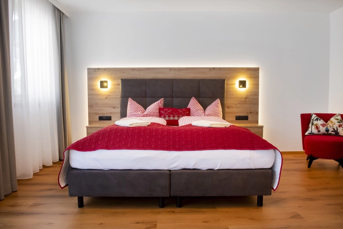 Modernes Doppelbett mit roter Decke und karierten Kissen, indirekte Beleuchtung und Holzrückwand, heller Raum mit Parkett.