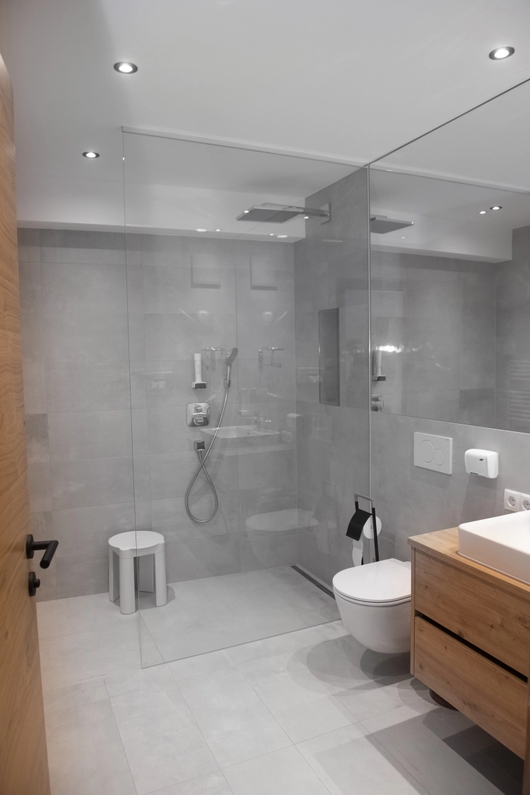 Modernes Bad in Grau mit bodengleicher Dusche, Holzwaschtisch, Hocker und großem Spiegel an der Wand.