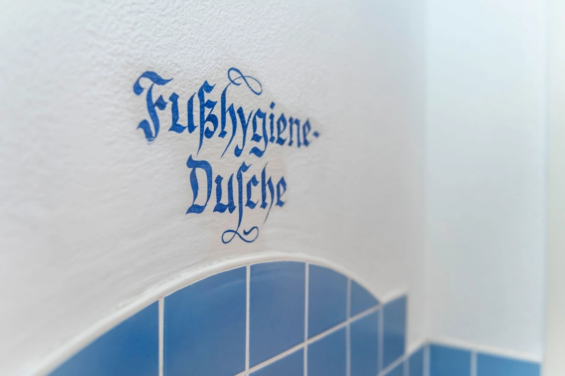 Das Bild zeigt den Schriftzug "Fußhygiene-Dusche" auf einer Wand über blauen Fliesen.