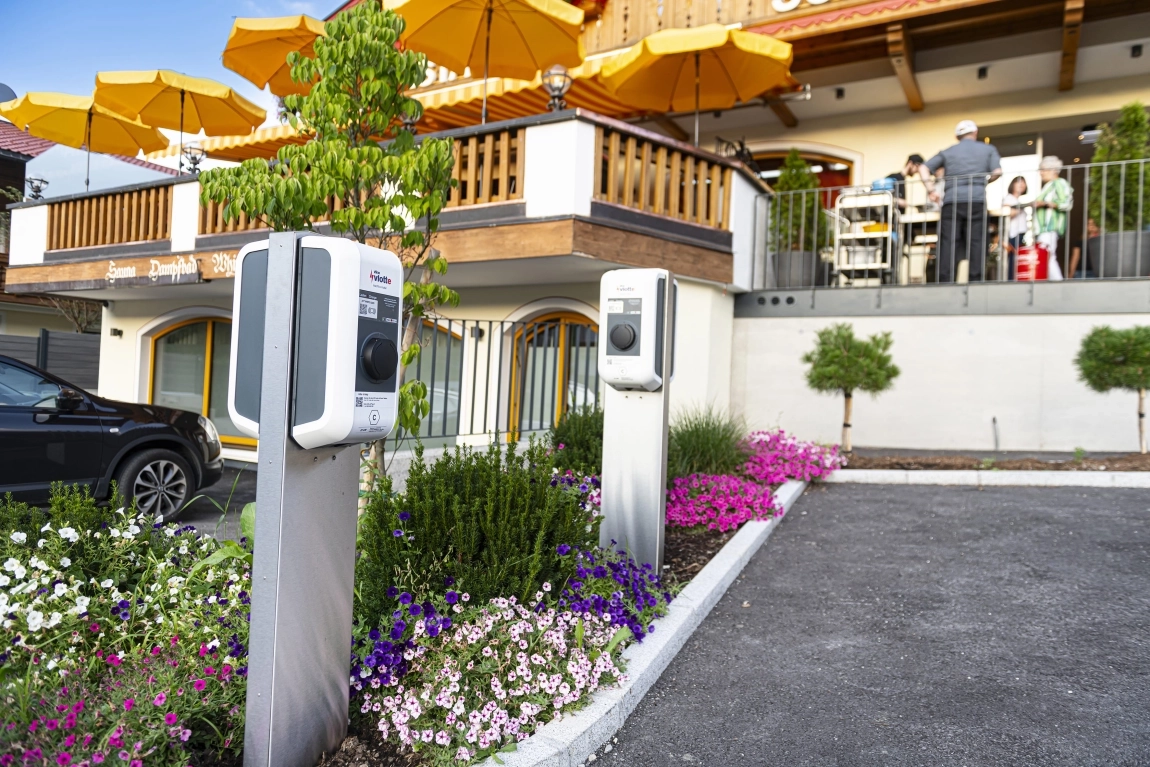Hotel mit Terrasse, Sonnenschirmen, zwei E-Ladestationen im Vorgarten mit Blumenbeet und angrenzendem Parkplatz.