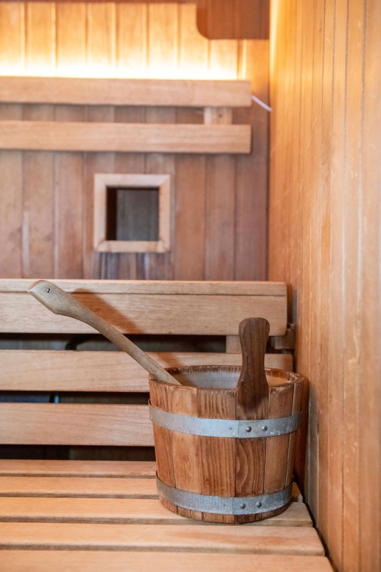 Holzaufgusskübel mit Kelle steht auf heller Holzbank in einer warm beleuchteten Sauna mit Holzverkleidung.