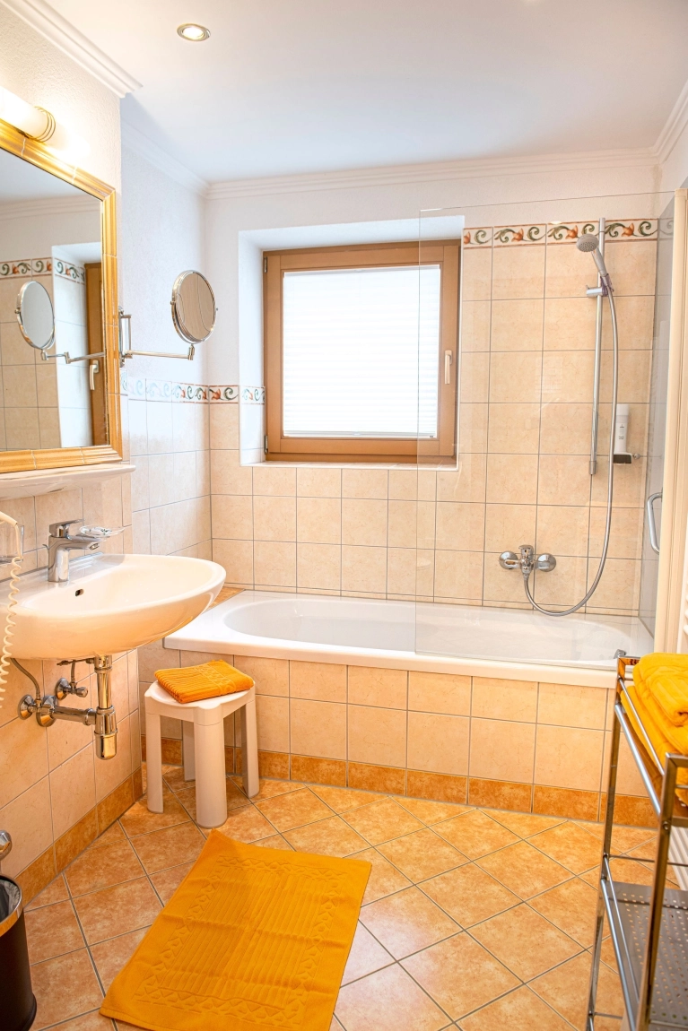Barrierefreies Bad mit Badewanne, Dusche, Waschbecken, Spiegel, Hocker und gelbem Teppich vor hellen Fliesen.