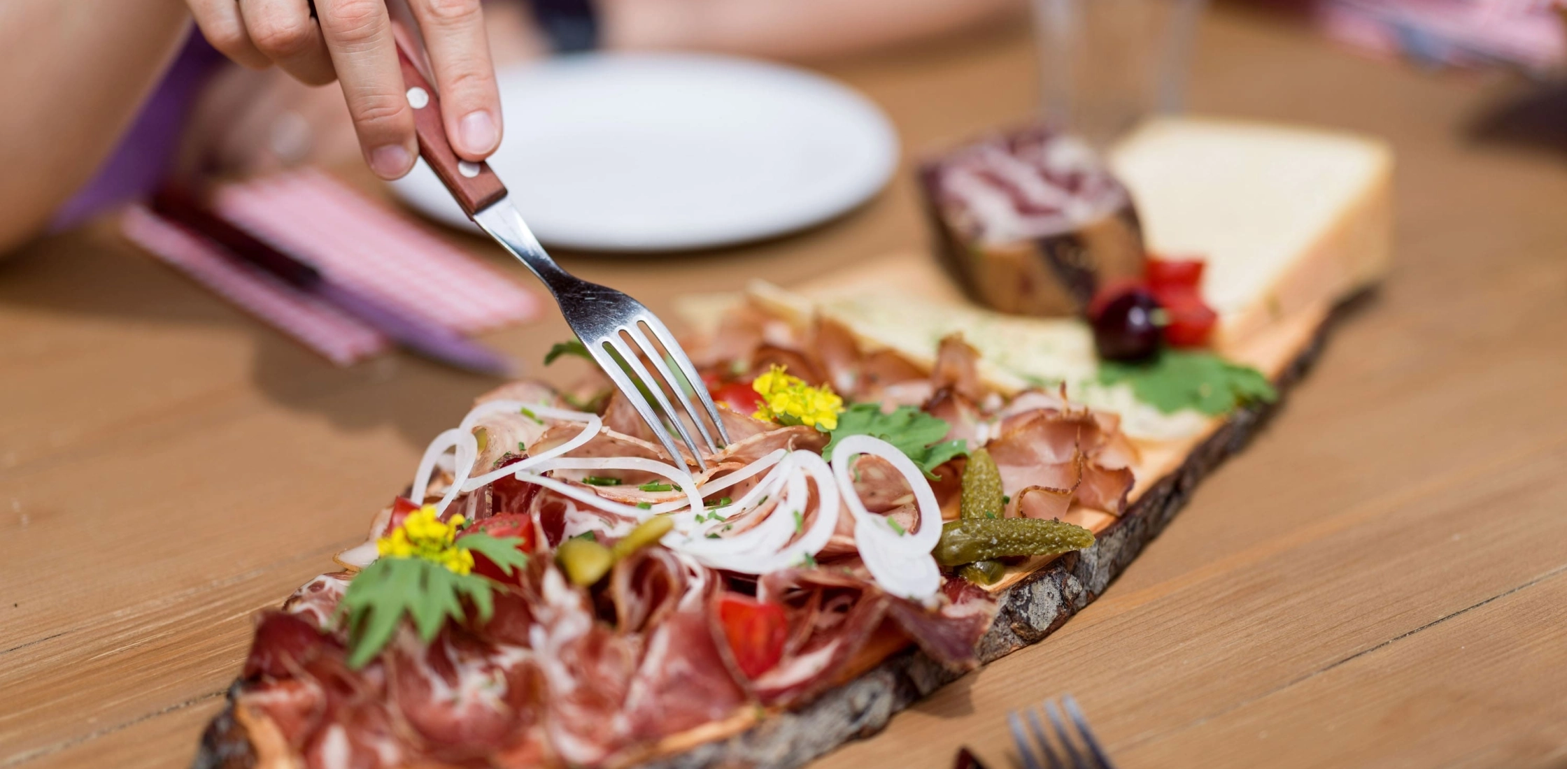 Eine Person isst eine Platte mit Schinken, Käse und Zwiebeln, verziert mit Kräutern und essbaren Blumen.