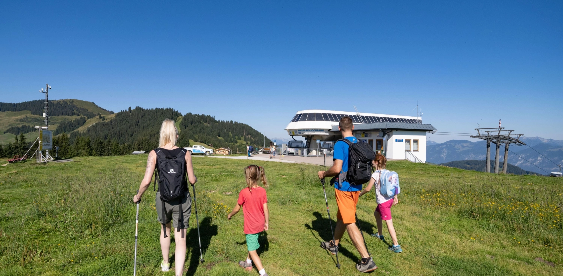 Eine Familie wandert auf einer grünen Wiese mit einer Gondelbahn im Hintergrund und einer malerischen Berglandschaft.