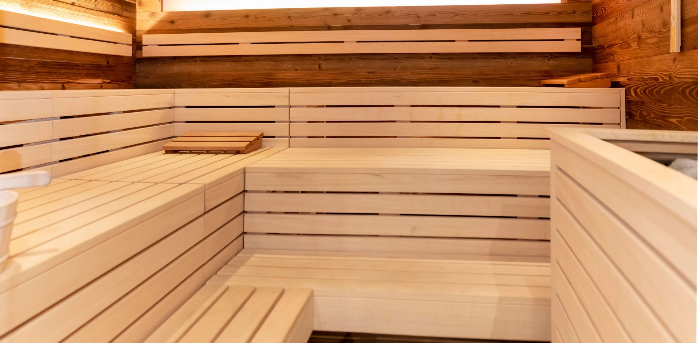 Innenansicht einer Sauna mit Holzbank und weichem, warmem Licht von einem Fenster. Eine gemütliche, entspannende Atmosphäre.