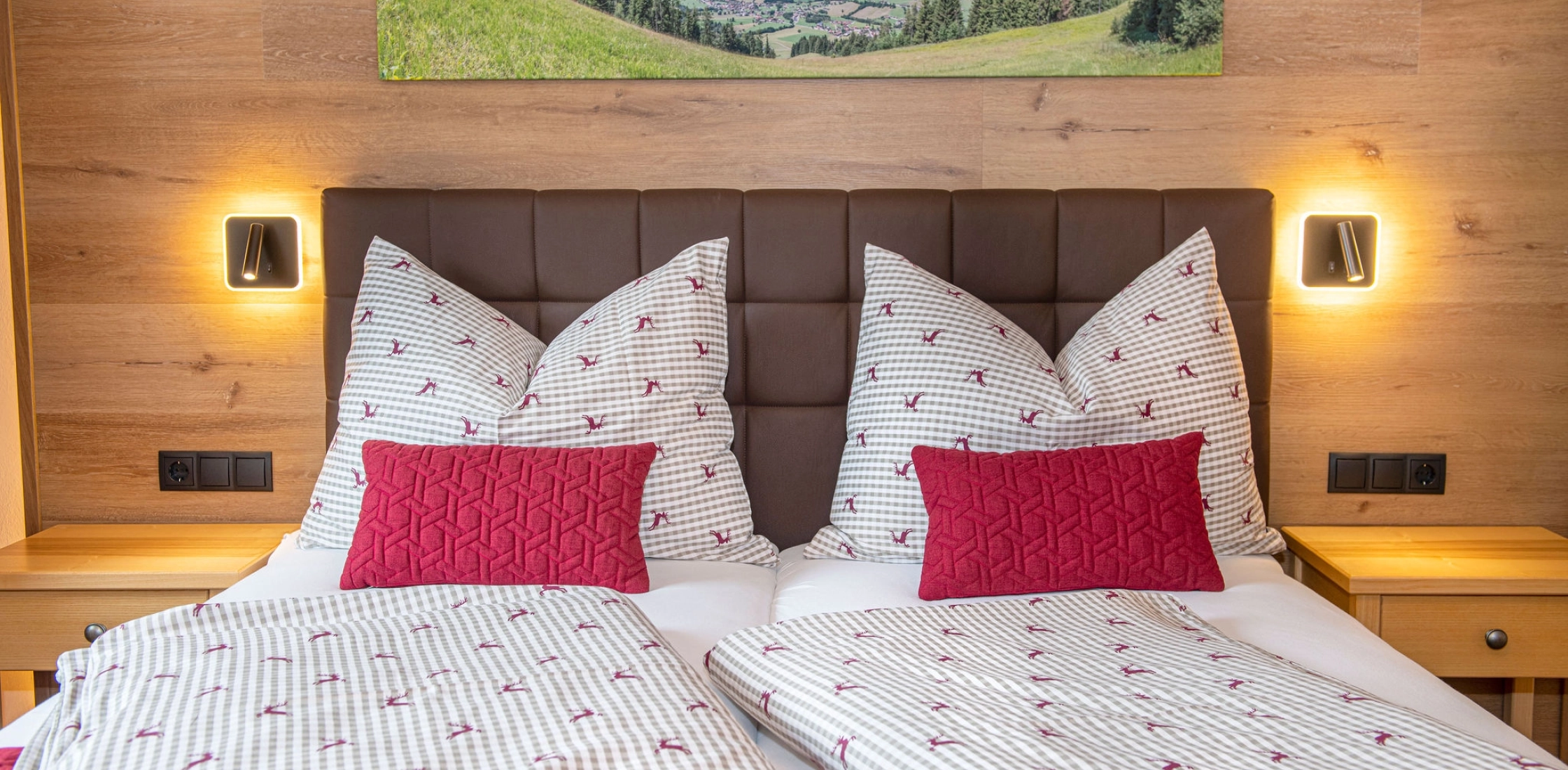 Helles Zimmer mit Doppelbett, roter Tagesdecke, Schreibtisch, TV an der Wand und Bergbild über dem Bett.