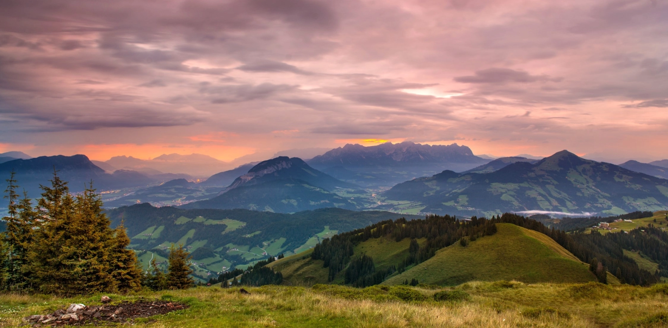 Berglandschaft bei Sonnenuntergang, mit sanften Hügeln, Bäumen und dramatischen Wolken am Himmel.