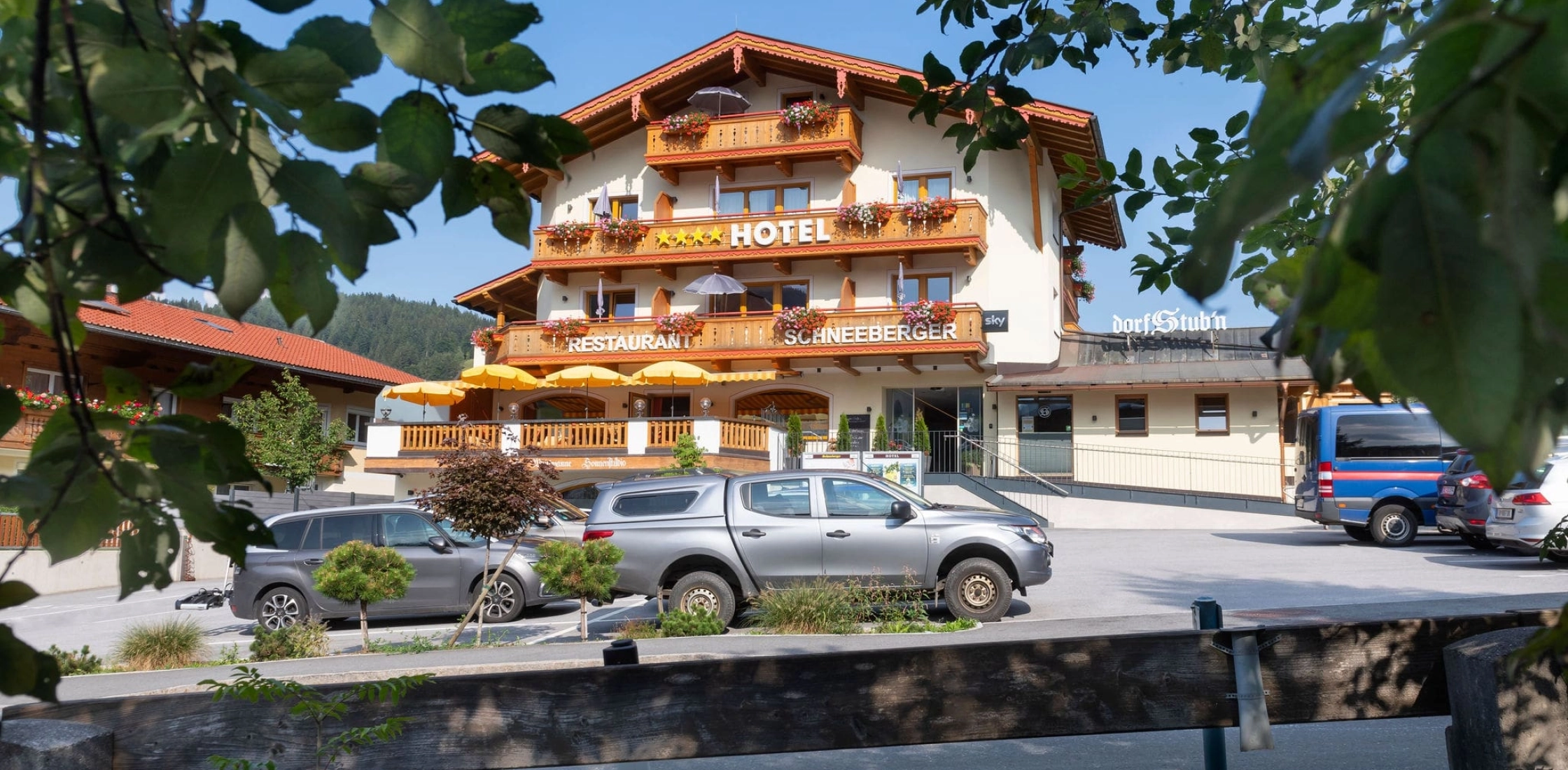 Frontansicht des Hotels Schneeberger mit Parkplatz, Terrasse, Rampe und Blick durch belaubte Äste im Vordergrund.