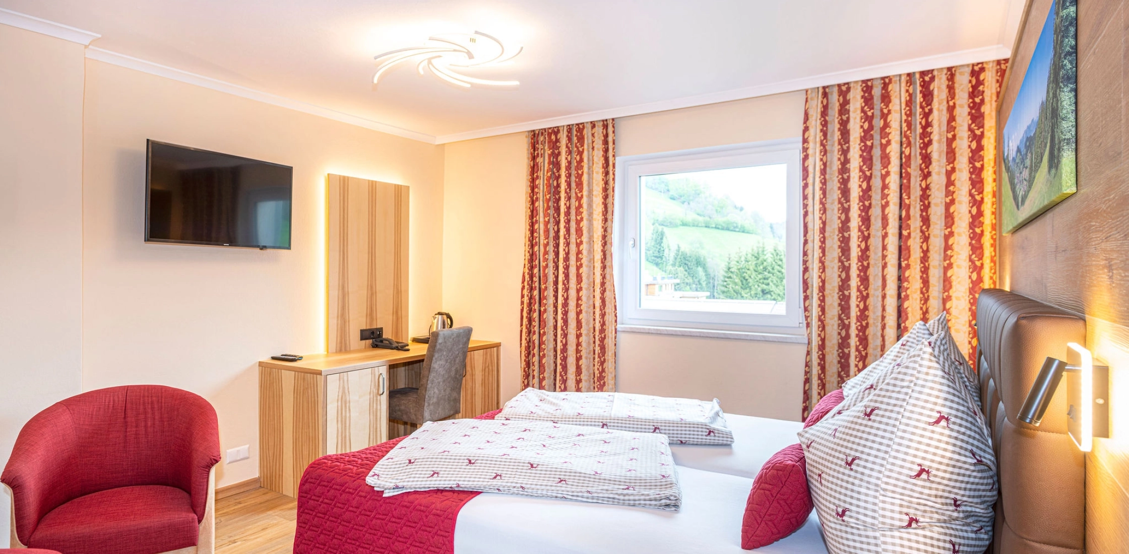 Helles Zimmer mit Doppelbett, roter Bettdecke, Schreibtisch, TV an der Wand, rotem Sessel und Fenster mit Ausblick.
