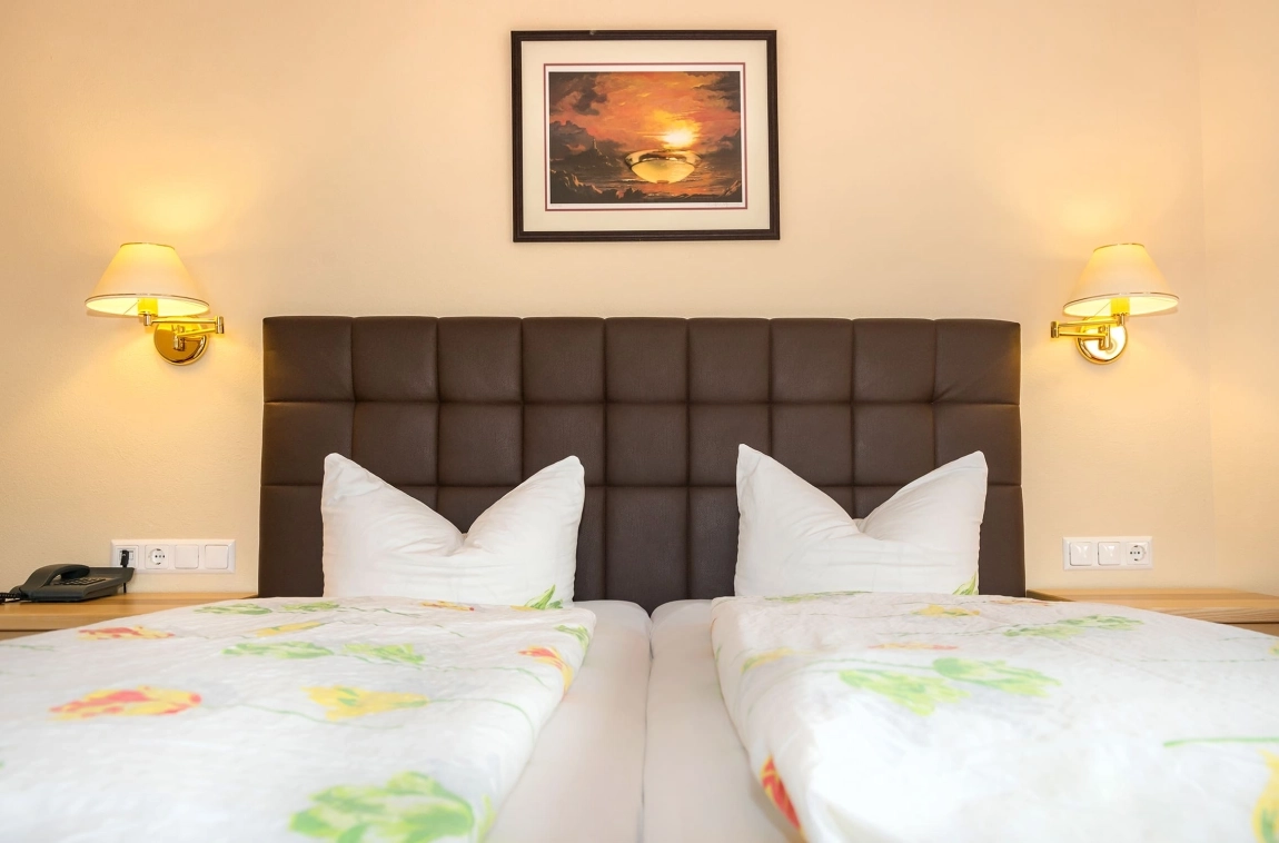 Doppelbett mit floralem Bettbezug, zwei Wandlampen und Sonnenuntergangsbild an der Wand über dem gepolsterten Kopfteil.