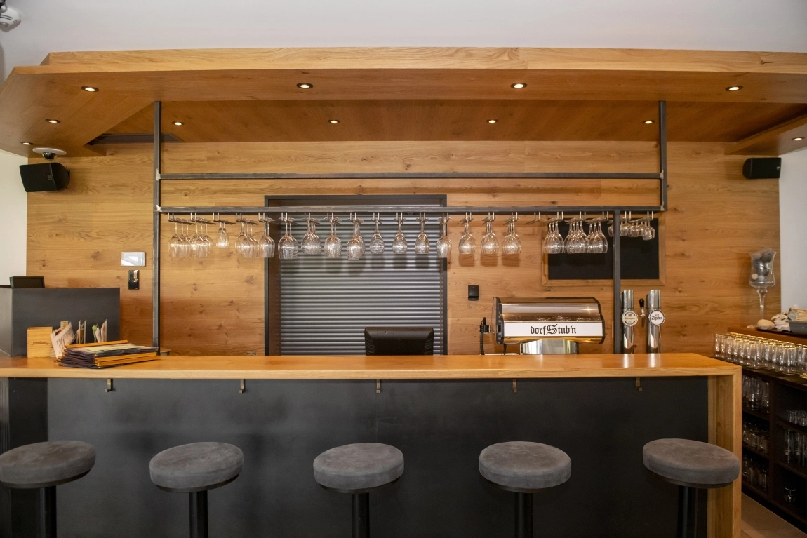 Moderne Bar der dorfStub’n bei Tageslicht: Stilvolles Holzdesign, klare Linien, Hocker mit grauen Polstern und bereit für den nächsten geselligen Abend.