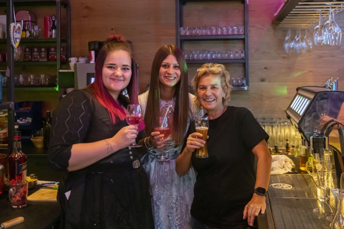 Drei Frauen des Teams der dorfStub’n stehen hinter der Bar, lachen und halten Getränke in der Hand.