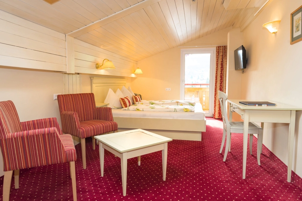 Helles Dachzimmer mit Doppelbett, TV, roten Sesseln, Tisch, Schreibtisch und Tür zum Balkon mit Tageslicht.