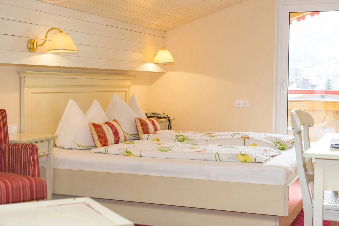 Gemütliches Doppelbett mit Blumenbettwäsche und Zierkissen, seitlich Schreibtisch und Zugang zum Balkon mit Holzbrüstung.