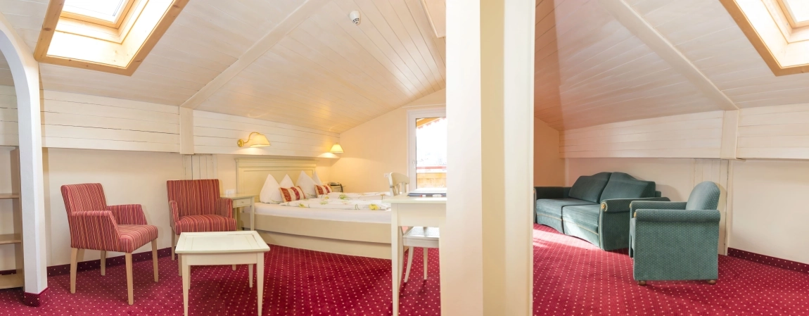 Helles Dachzimmer mit Dachfenstern, Doppelbett, Sitzecke, rotem Teppich und grünem Sofa unter schräger Decke.