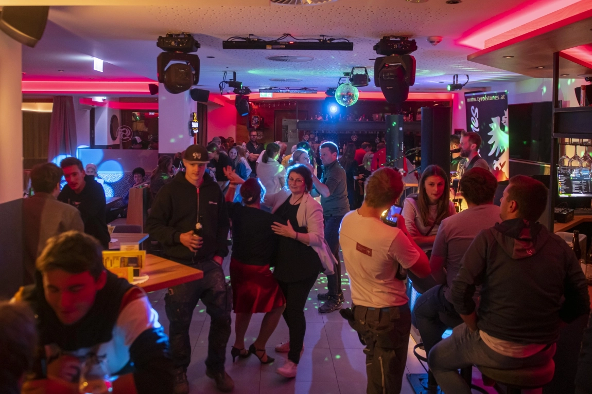Abendstimmung in der Bar mit roten Lichtern, tanzenden Gästen, Gesprächen an der Theke und Discokugel.