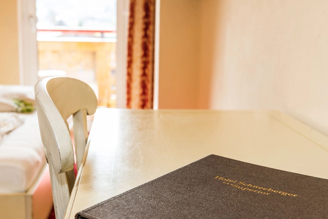 Heller Schreibtisch mit Stuhl nahe einem Bett, darauf eine schwarze Infomappe, im Hintergrund Zugang zum Balkon.
